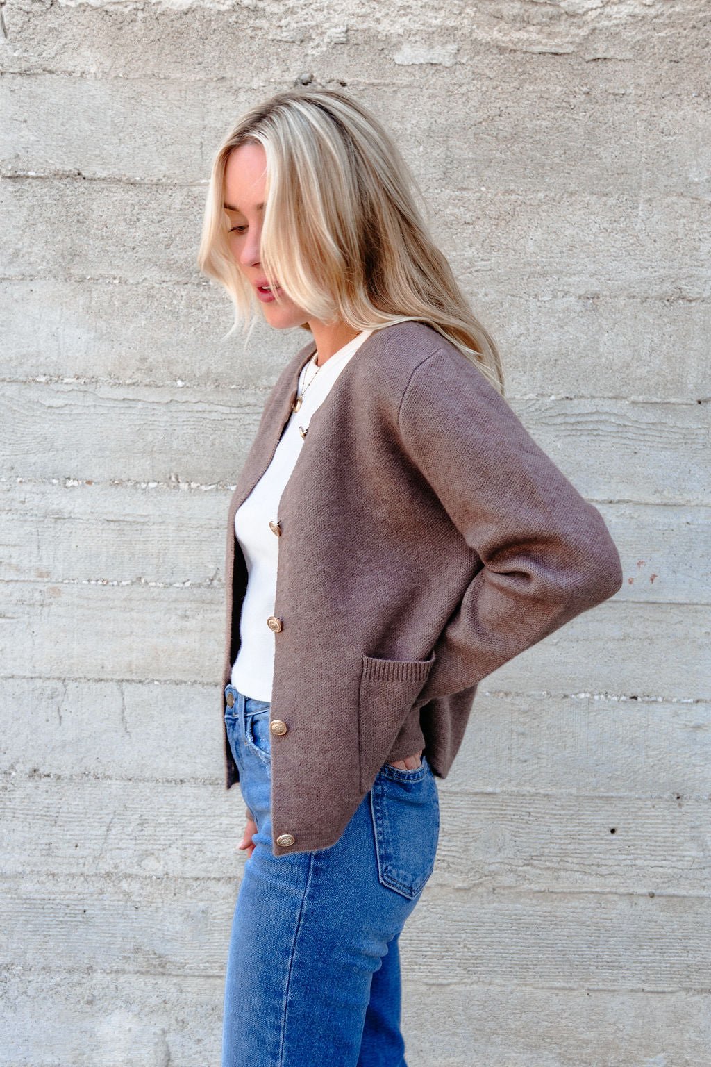 Woman with blonde hair wears the Autumn Button Front Sweater Cardigan in brown, white top, and blue jeans by a concrete wall.