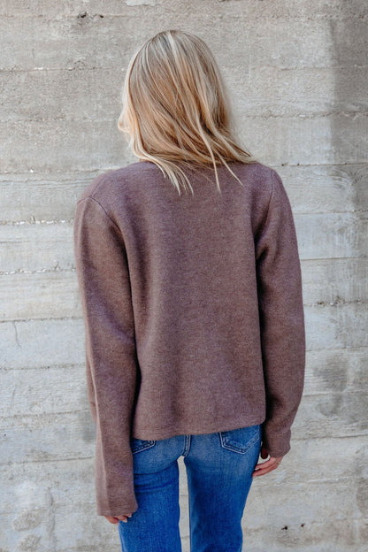A person with blonde hair wears the Autumn Button Front Sweater Cardigan - Brown and blue jeans, facing a concrete wall.