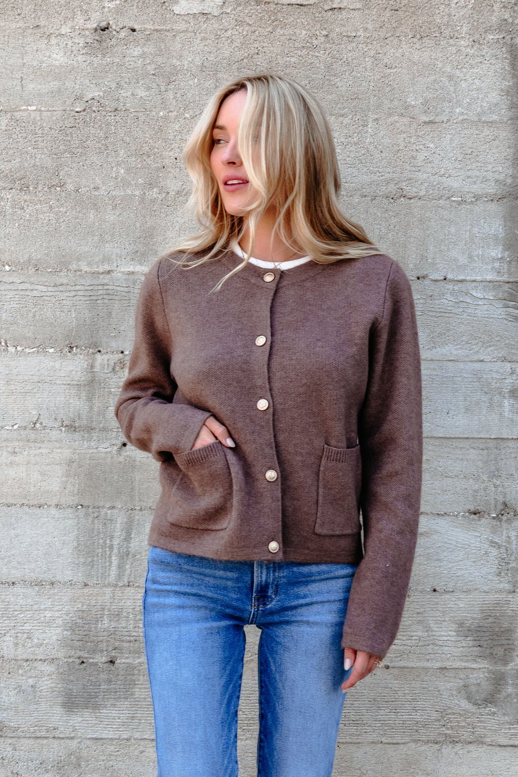 Woman wears the Autumn Button Front Sweater Cardigan in brown with gold buttons and blue jeans, standing by a concrete wall.