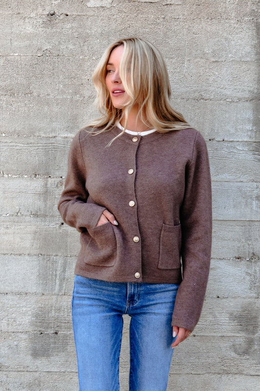 Woman wears the Autumn Button Front Sweater Cardigan in brown with gold buttons and blue jeans, standing by a concrete wall.
