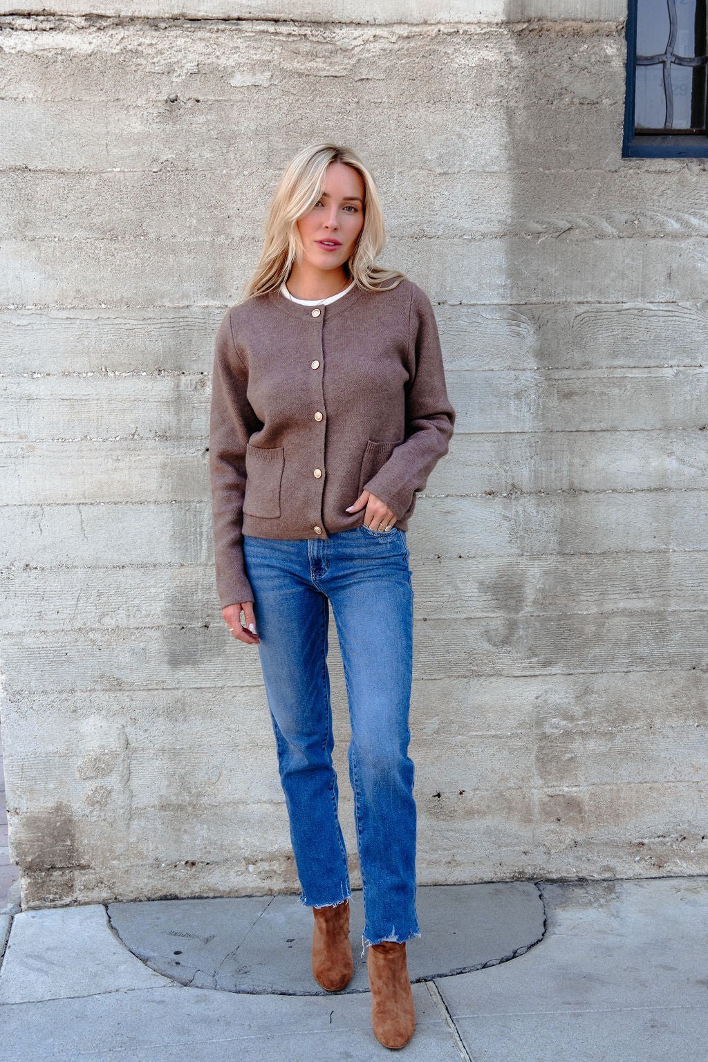 Woman stands against a concrete wall wearing the Autumn Button Front Sweater Cardigan - Brown, blue jeans, boots, and a pearl necklace.