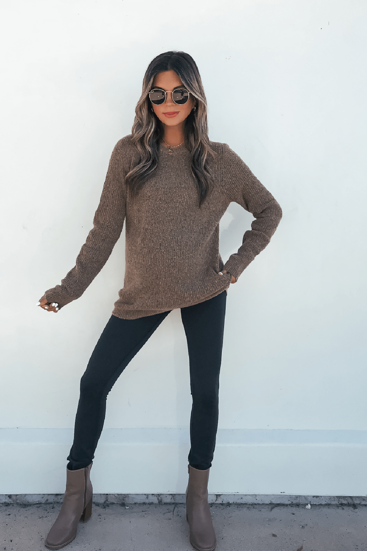 Woman in sunglasses wears the Autumn Glow Brushed Ochre Waffle Knit Sweater with black leggings and brown boots against a white wall.