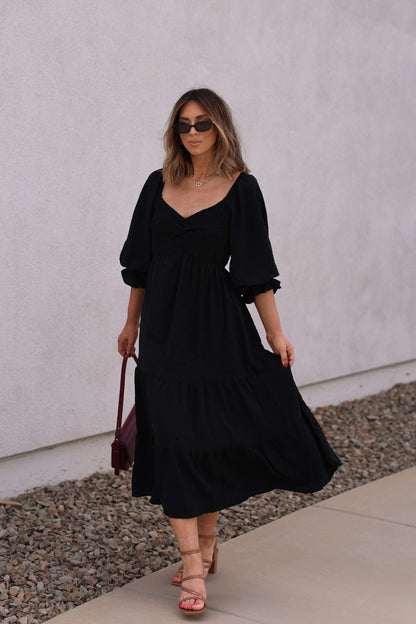 Woman in the Autumn Nights Black Tiered Midi Dress walks along a sidewalk by a white wall, wearing sunglasses and sandals.