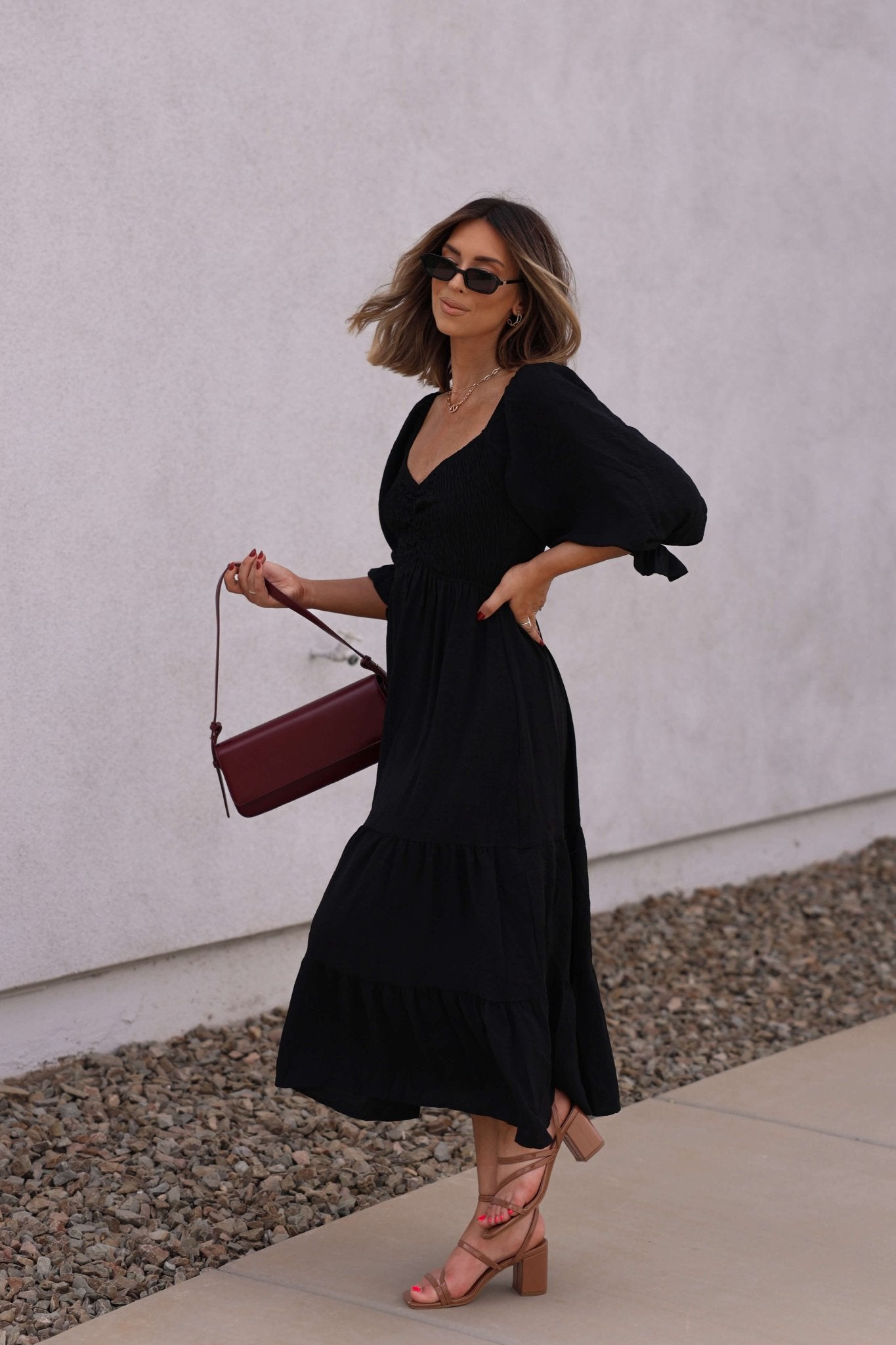 Woman in the Autumn Nights Black Tiered Midi Dress walks outside, carrying a burgundy handbag and wearing tan block-heeled sandals.