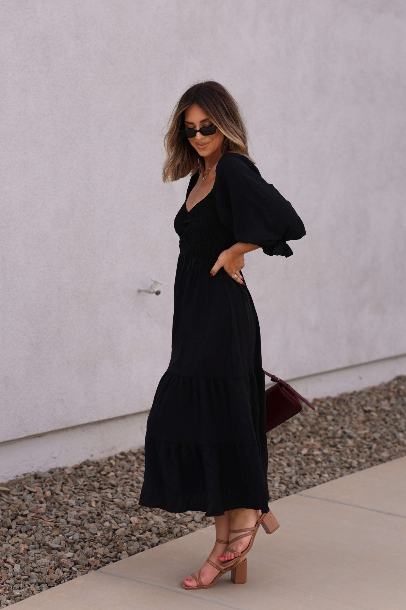 Woman in the Autumn Nights Black Tiered Midi Dress stands on a sidewalk, wearing sunglasses, heeled sandals, and holding a bag.