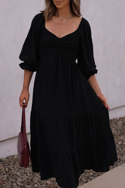 A woman in the Autumn Nights Black Tiered Midi Dress stands on a sidewalk near a white wall, holding a burgundy handbag.