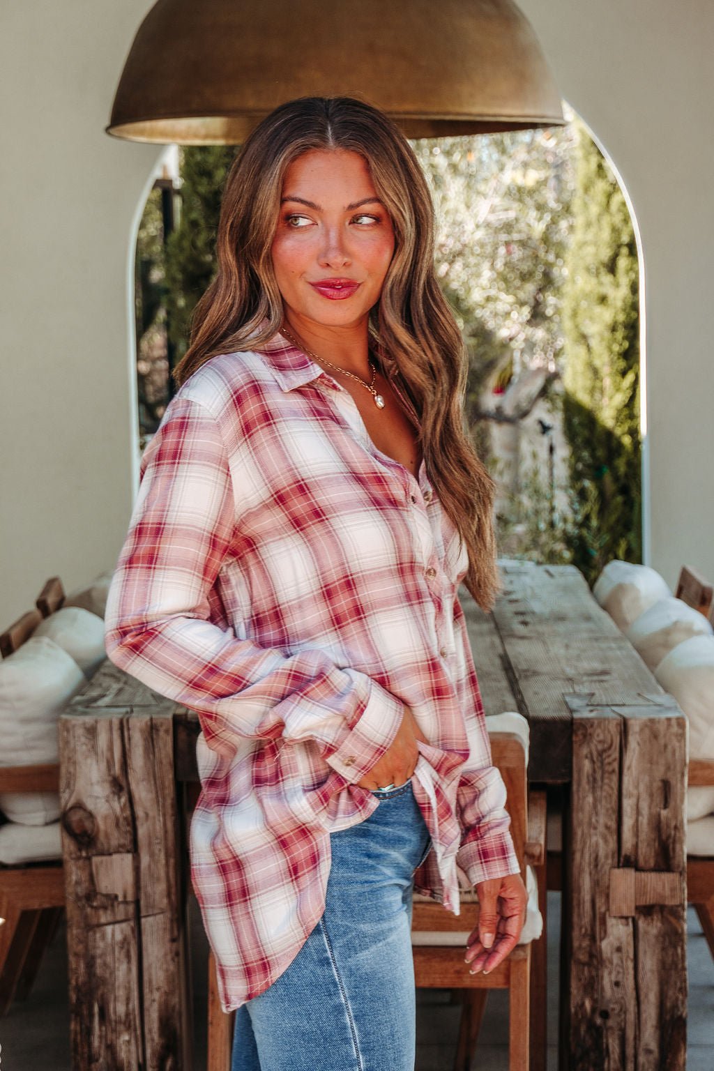 Woman with long wavy hair in an Autumn Spice Wine Plaid Shirt and jeans stands by a rustic wooden table, smiling over her shoulder.