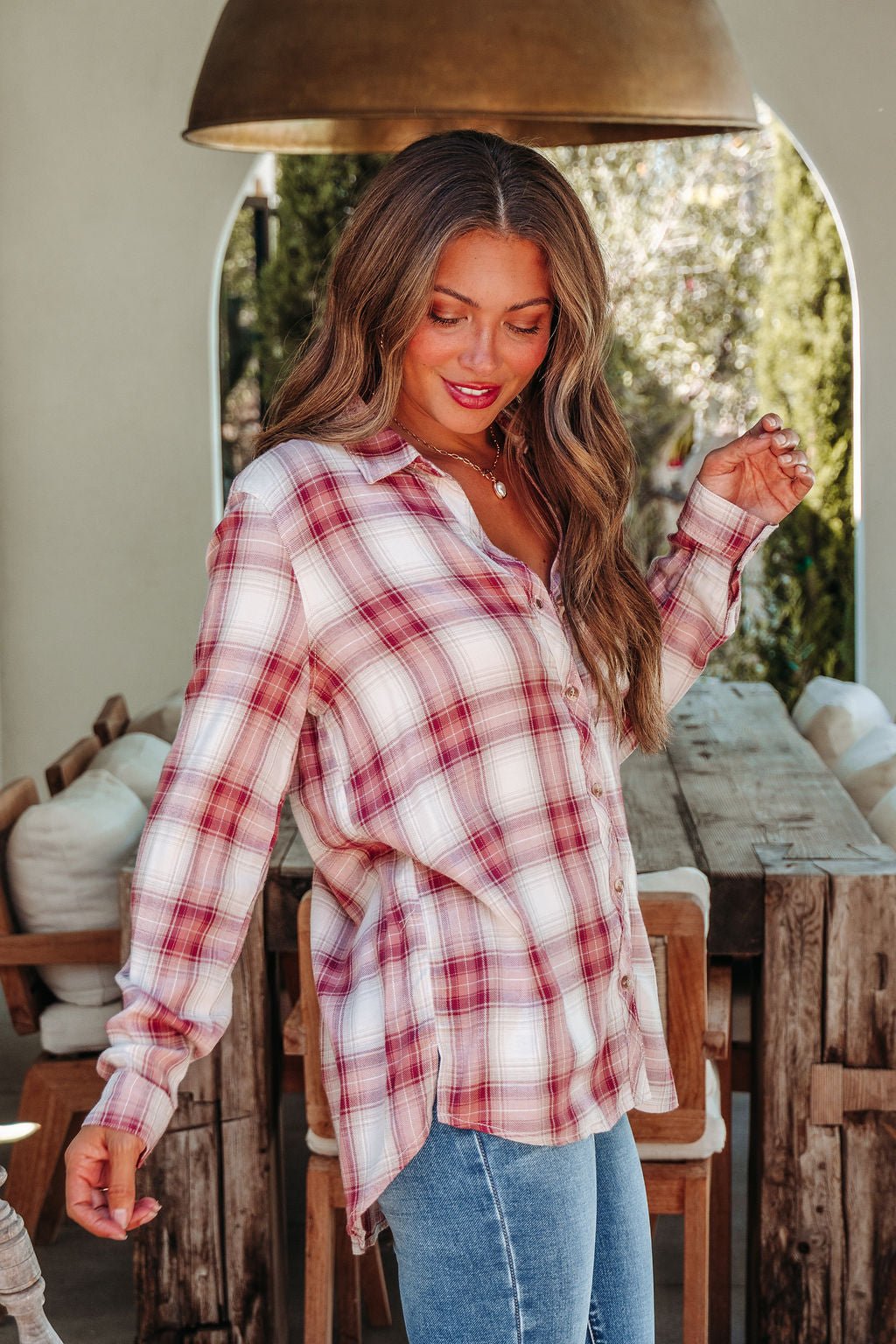 A woman in an Autumn Spice Wine Plaid Shirt and blue jeans stands indoors, smiling warmly with one hand raised.