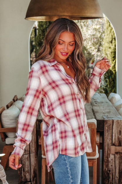 A woman in an Autumn Spice Wine Plaid Shirt and blue jeans stands indoors, smiling warmly with one hand raised.