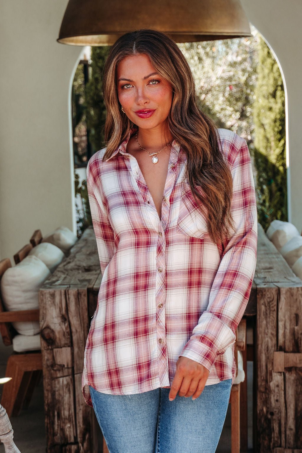 A woman in an Autumn Spice Wine Plaid Shirt and jeans stands by a rustic wooden table, smiling softly at the camera.