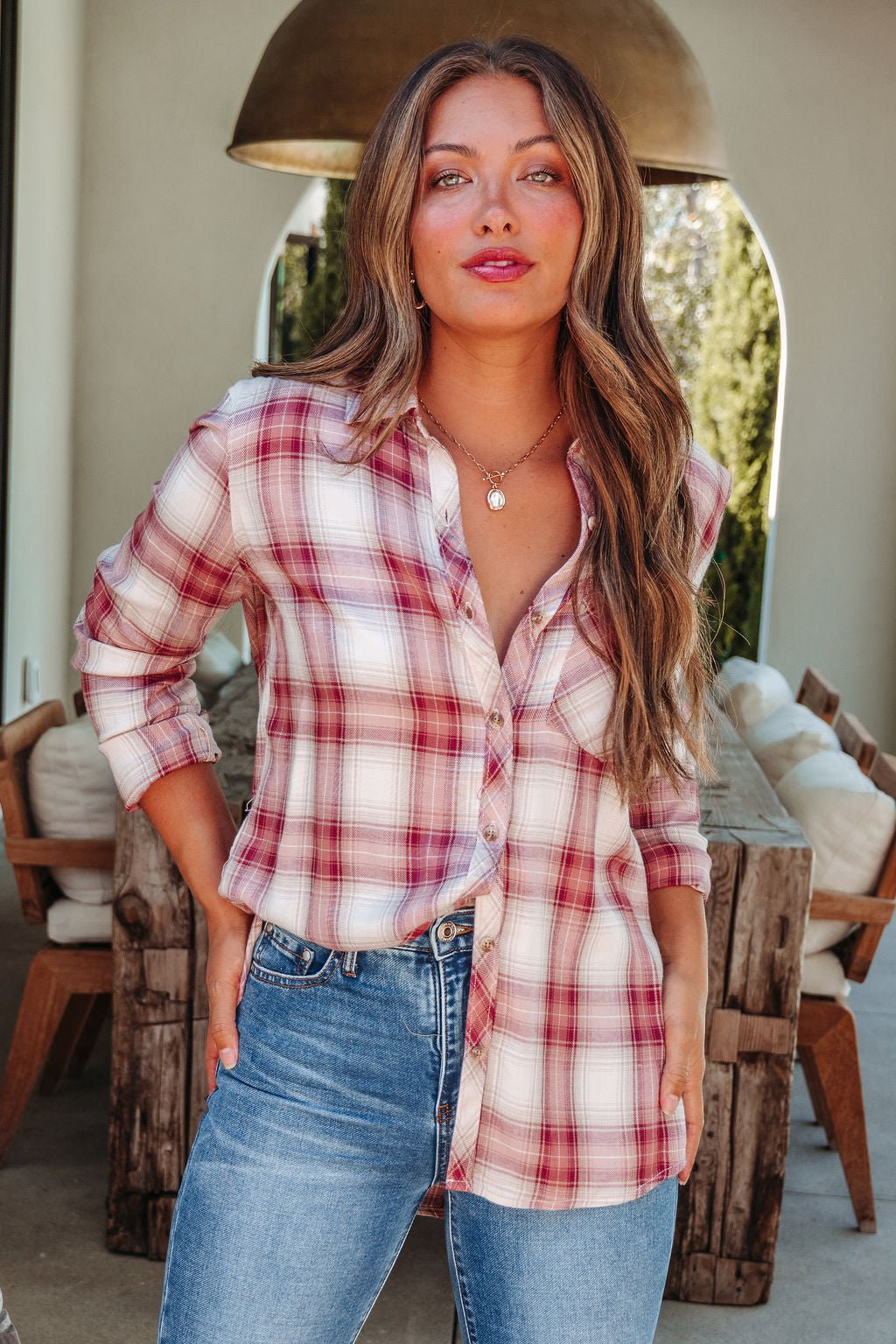 A woman with long brown hair wears an Autumn Spice Wine Plaid Shirt and blue jeans, standing indoors by a rustic wooden table.
