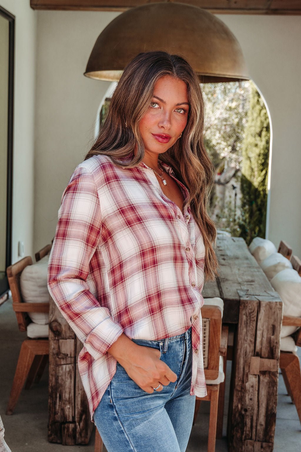 Woman with long wavy hair in an Autumn Spice Wine Plaid Shirt stands by a rustic wooden table, enhancing her fall style.