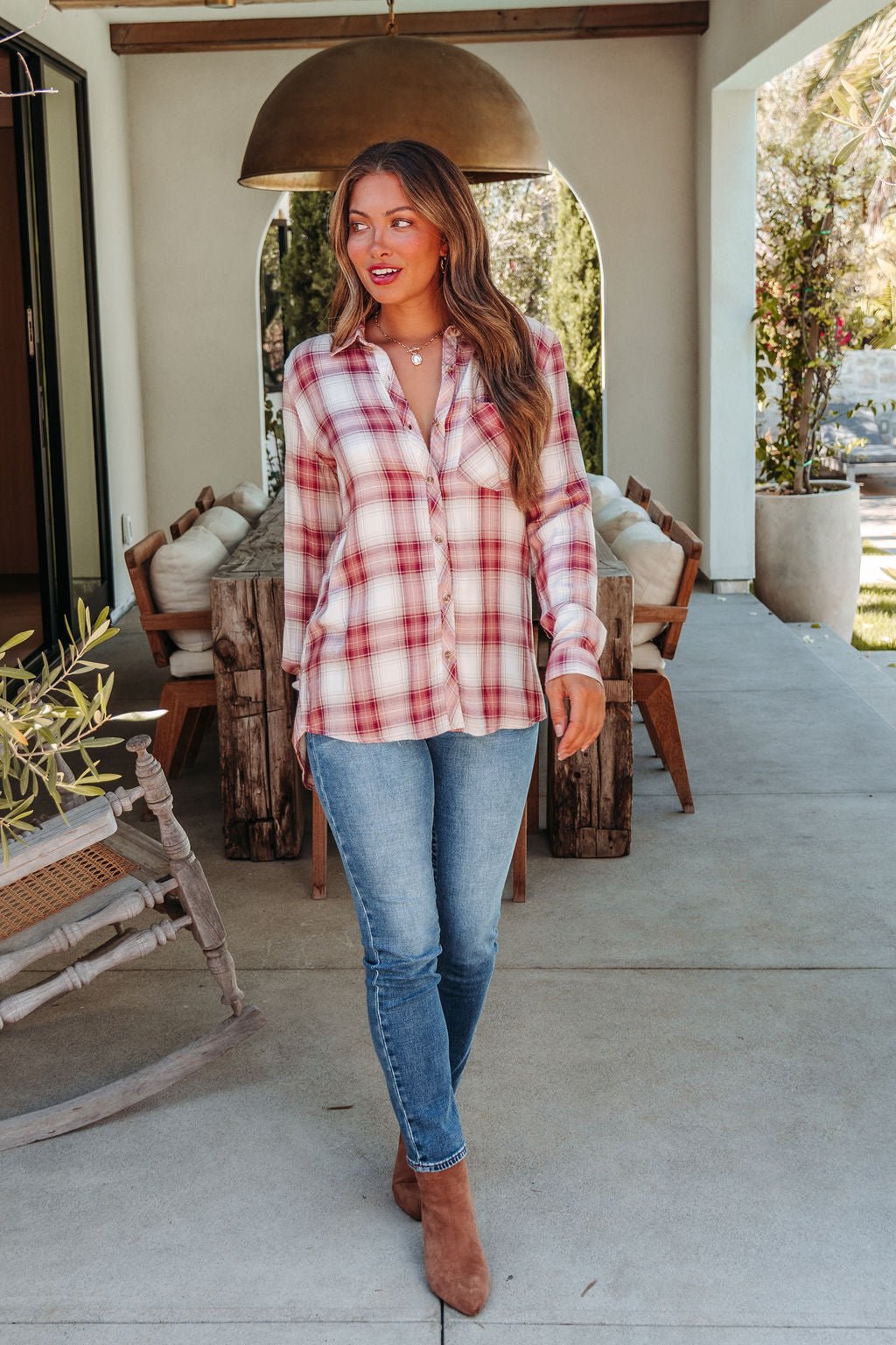 A woman in an Autumn Spice Wine Plaid Shirt, blue jeans, and brown boots walks by a wooden patio dining table outdoors.