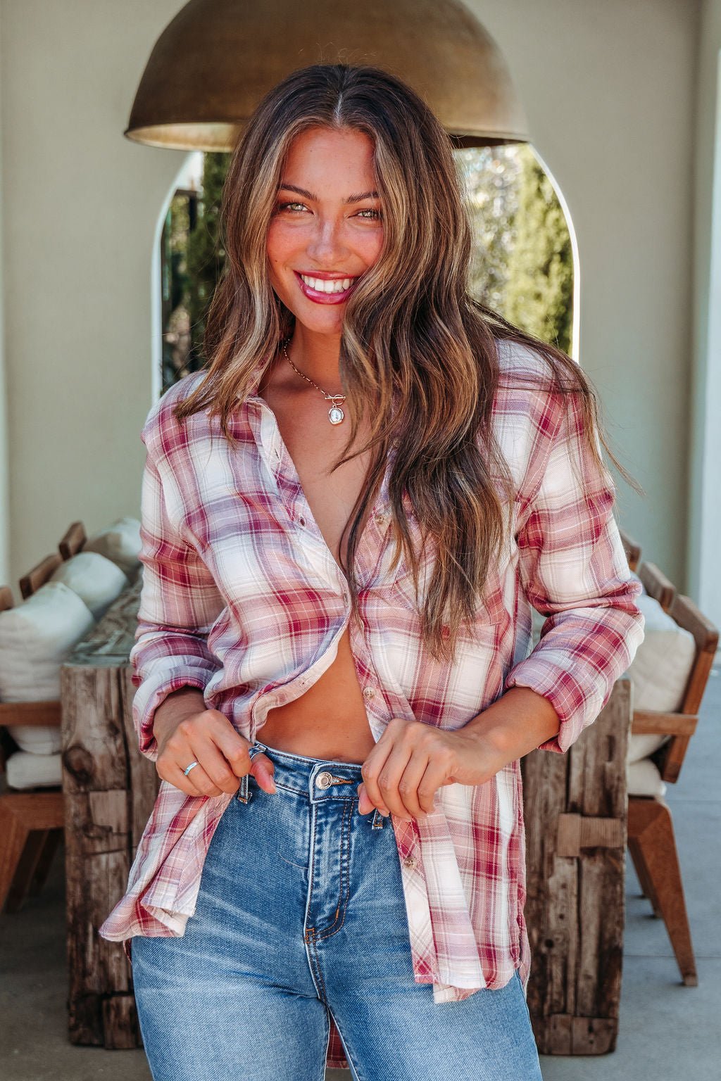 Smiling woman in an Autumn Spice Wine Plaid Shirt and blue jeans stands indoors, holding the front of her shirt open.