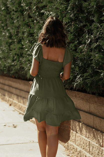 A woman in Ava's Sweetheart Olive Mini Dress walks along a sunlit sidewalk next to a green hedge and brick wall.
