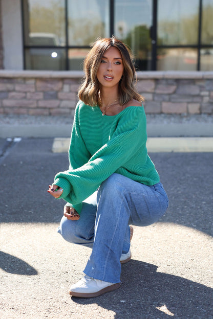 Woman with shoulder-length hair wears the Avery Ribbed Boat Neck Sweater in green while squatting outdoors on pavement.