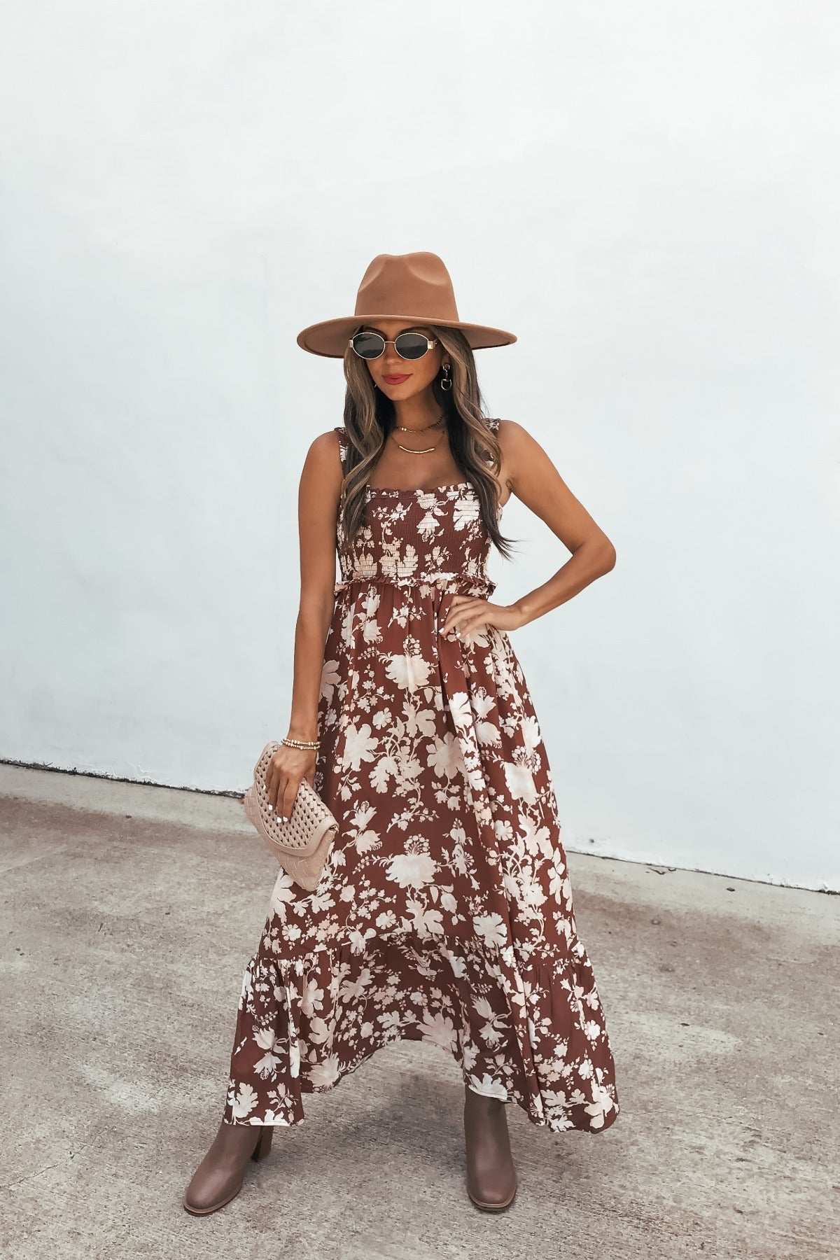 Woman in the Azalea Floral Maxi Dress - Brown, with a brown hat, boots, sunglasses, and beige clutch stands on concrete by a white backdrop.