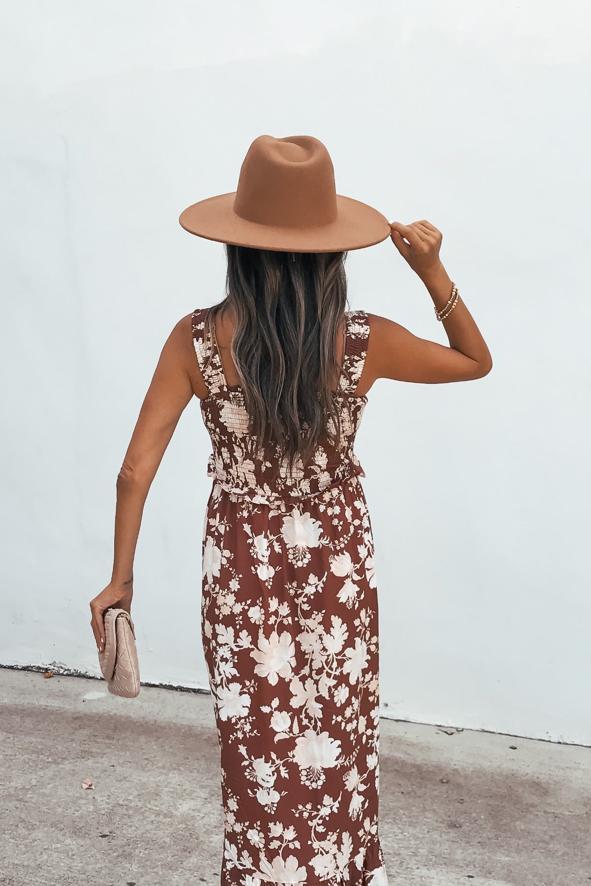 Woman in the Azalea Floral Maxi Dress - Brown and wide-brim hat stands by a white wall, holding a clutch.