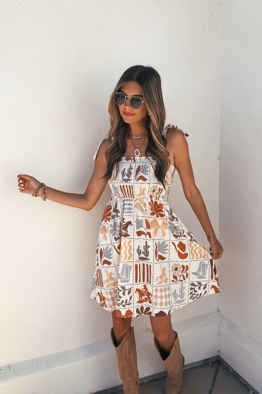 Woman wears the Baevely Natural Print Mini Dress, featuring a smocked bodice and tie straps, standing by a white wall.