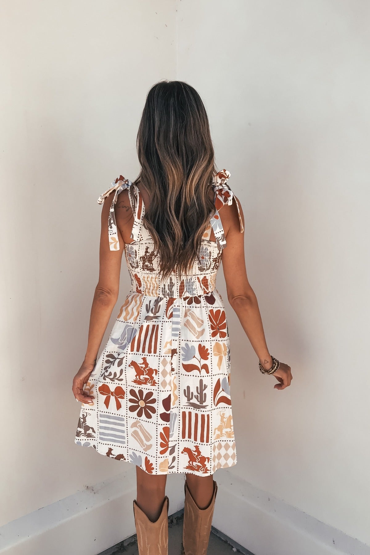 Woman with long wavy hair in the Baevely Natural Print Mini Dress and cowboy boots, standing against a white wall, facing away.