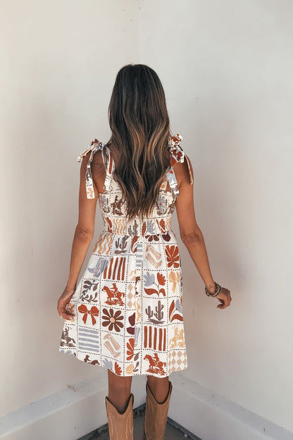 Woman with long wavy hair in the Baevely Natural Print Mini Dress and cowboy boots, standing against a white wall, facing away.