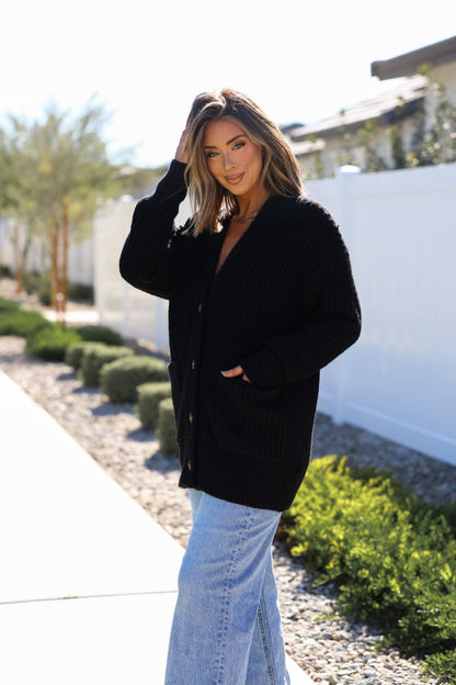 Woman wears a Basic Black Knit Button Down Cardigan and light blue jeans outdoors, smiling as she touches her hair.