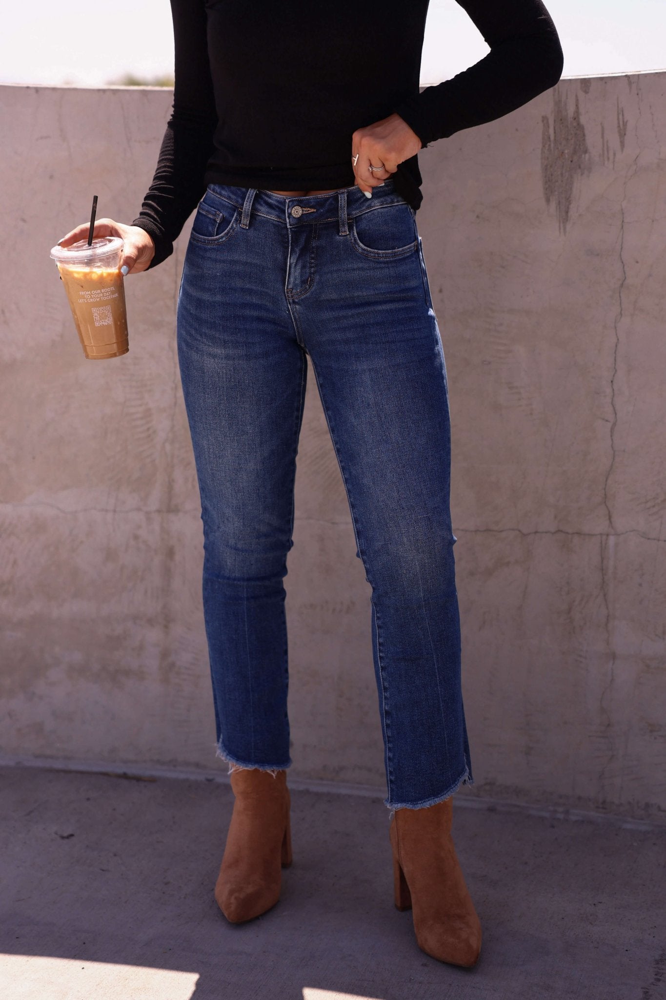 Person in a black long-sleeve top, Bayeas Dark Wash Mid Rise Bootcut Jeans, tan boots, holding iced coffee by a concrete wall.