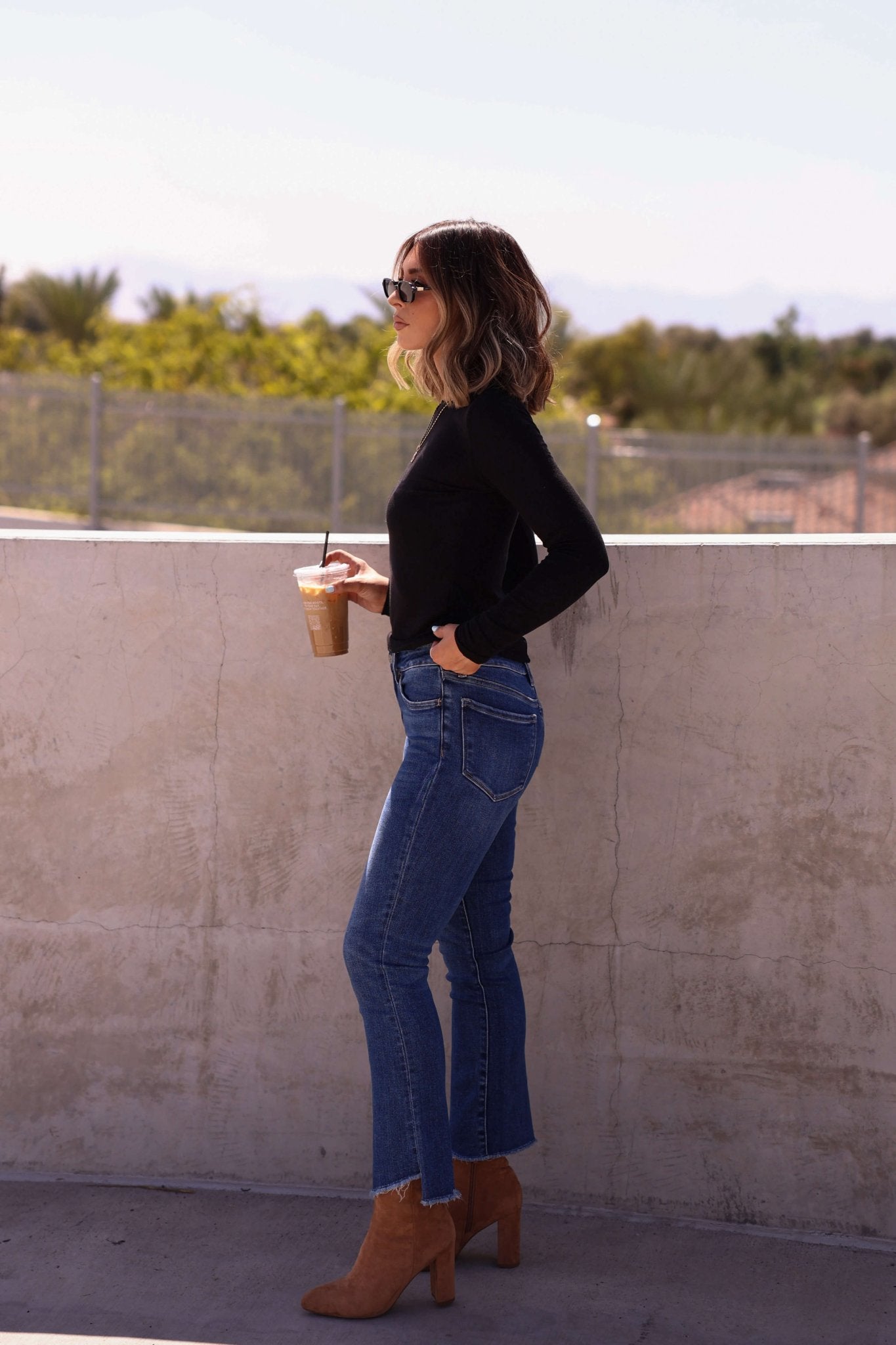 Woman in sunglasses, black top, and Bayeas Dark Wash Mid Rise Bootcut Jeans holding iced coffee by a concrete wall outdoors.