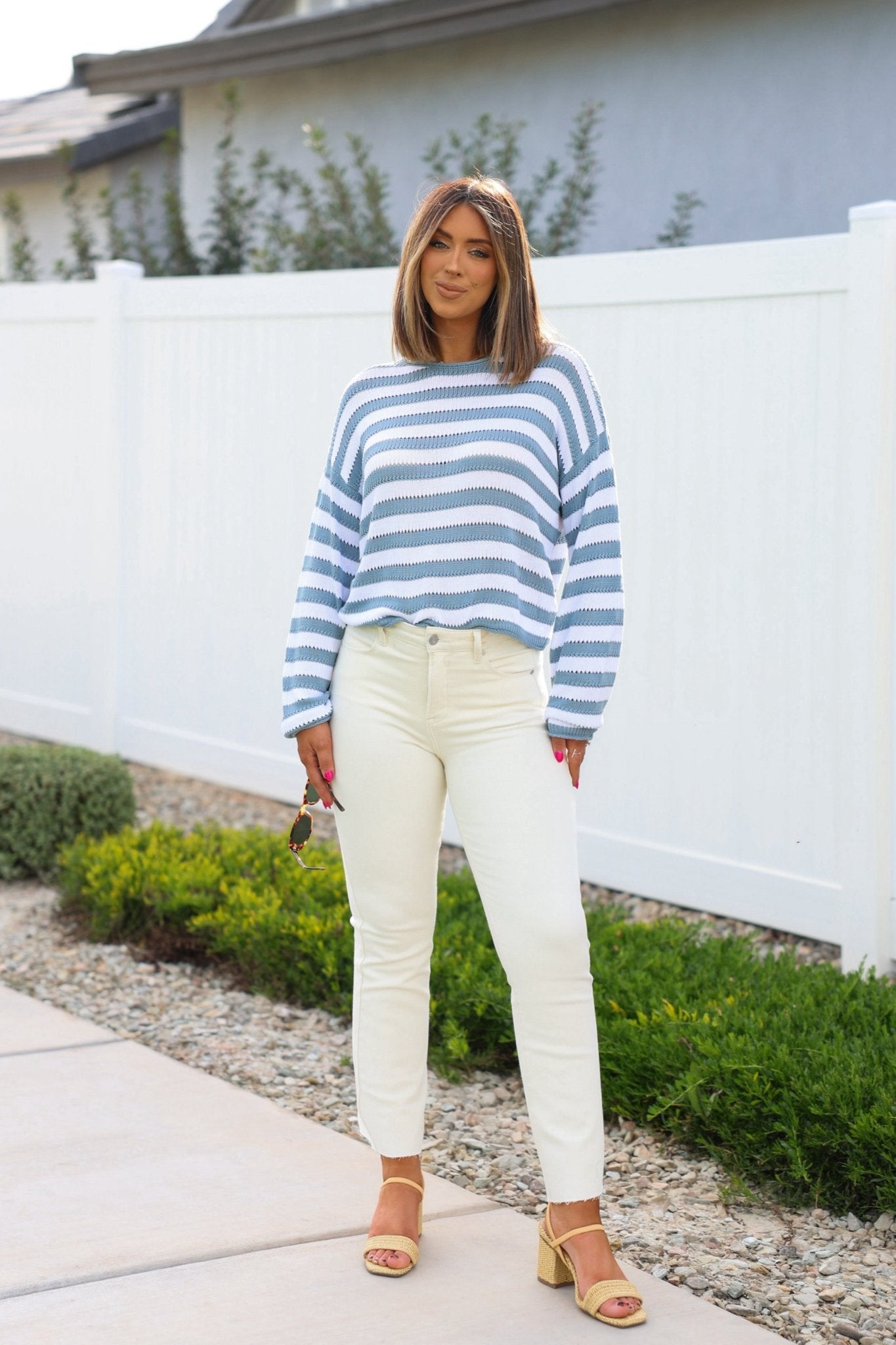 A woman stands outdoors in a striped sweater, beige sandals, and Bayeas Ivory Mid Rise Straight Leg Jeans, holding sunglasses.