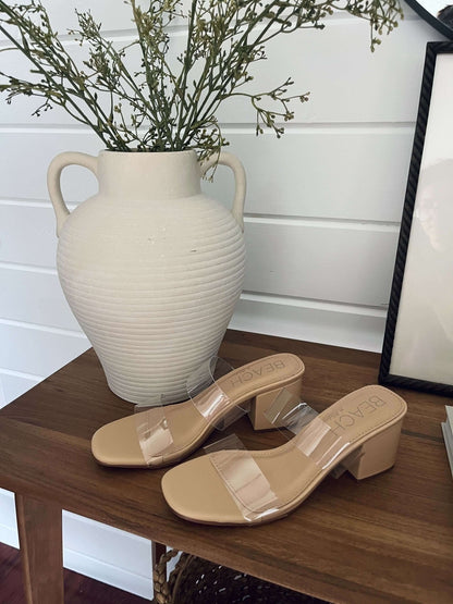 Beach by Matisse Aura Nude Strappy Heels with clear straps sit on a wooden table next to a white ceramic vase with greenery.