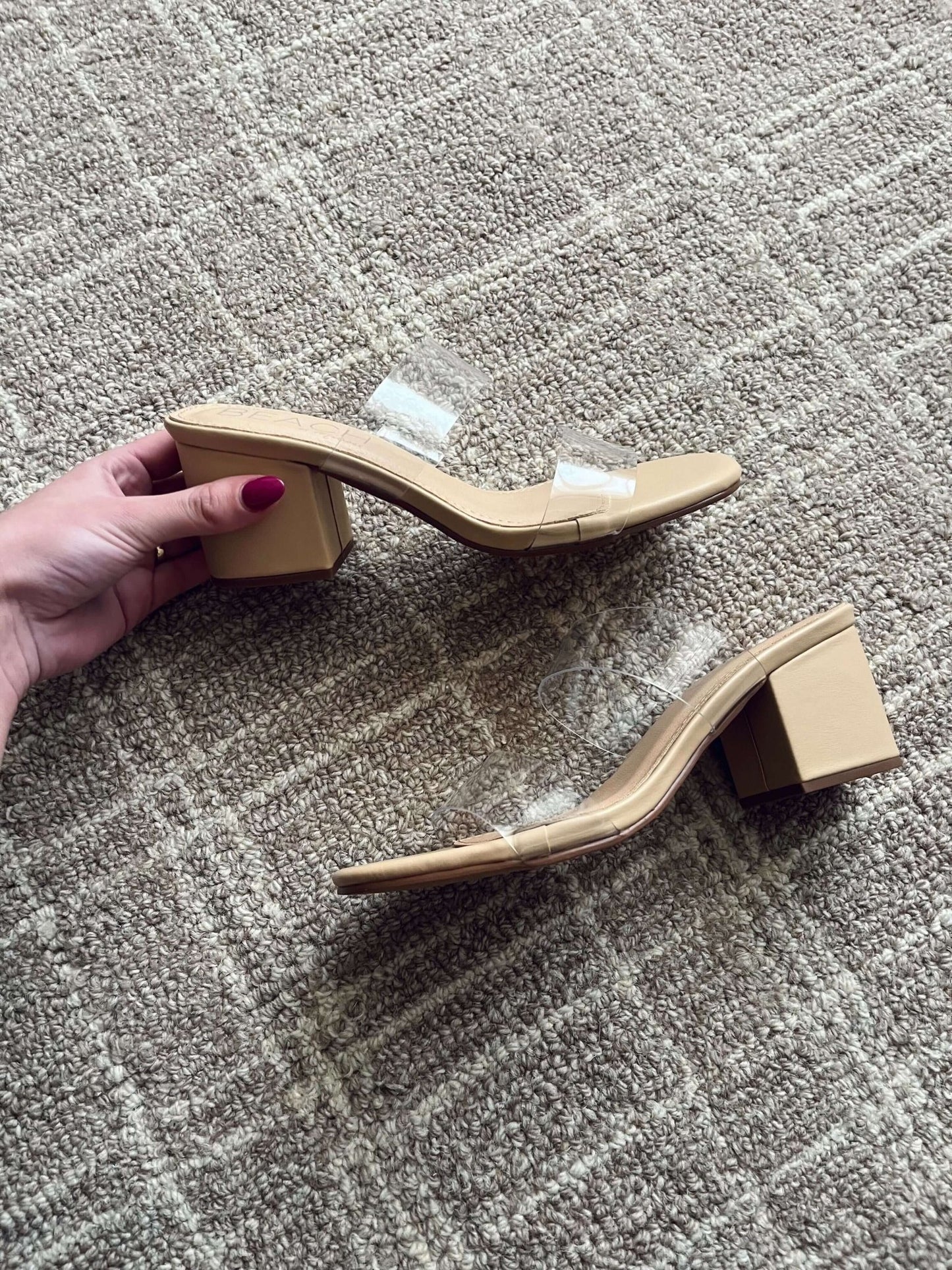 A hand holds a Beach by Matisse Aura Nude Strappy Heel with clear straps, next to its pair on a textured carpet.