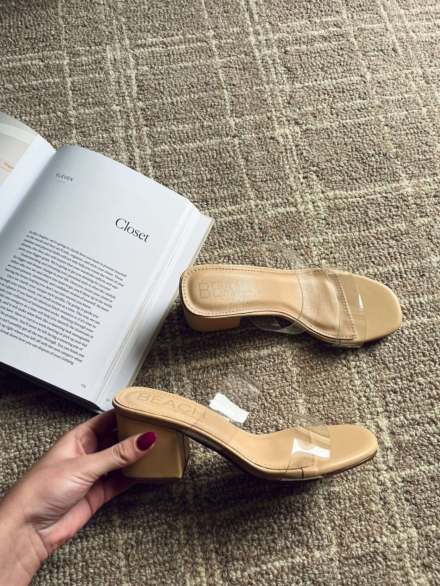 A hand holds Beach by Matisse Aura Nude Strappy Heels with clear straps beside an open book and matching sandal on a tan carpet.