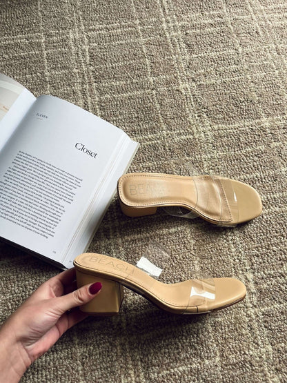 A hand holds Beach by Matisse Aura Nude Strappy Heels with clear straps beside an open book and matching sandal on a tan carpet.