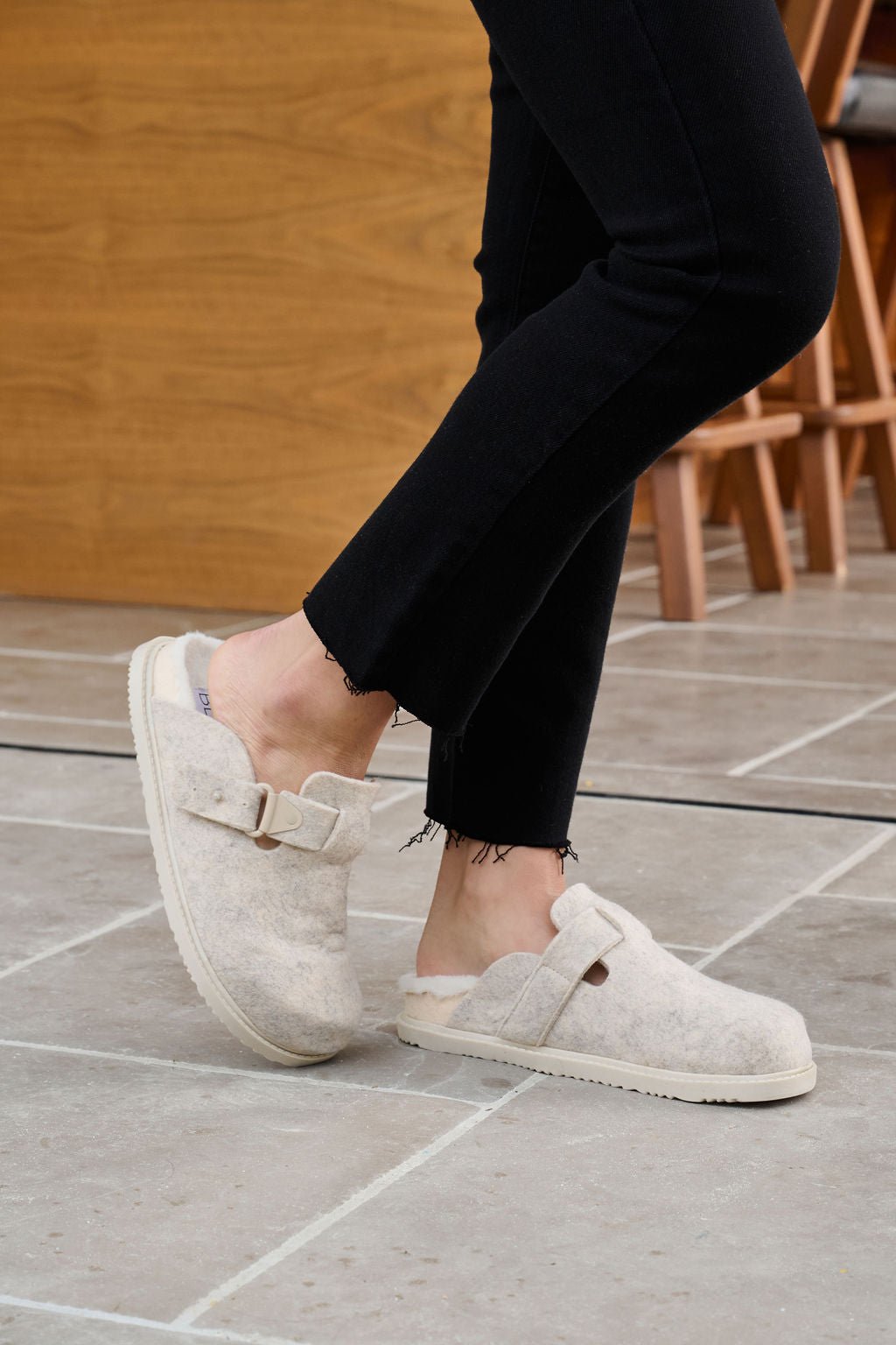 Wearing Beach By Matisse Portland Oatmeal Brushed Clogs and black pants, a person stands on tile with a wooden chair in the background.