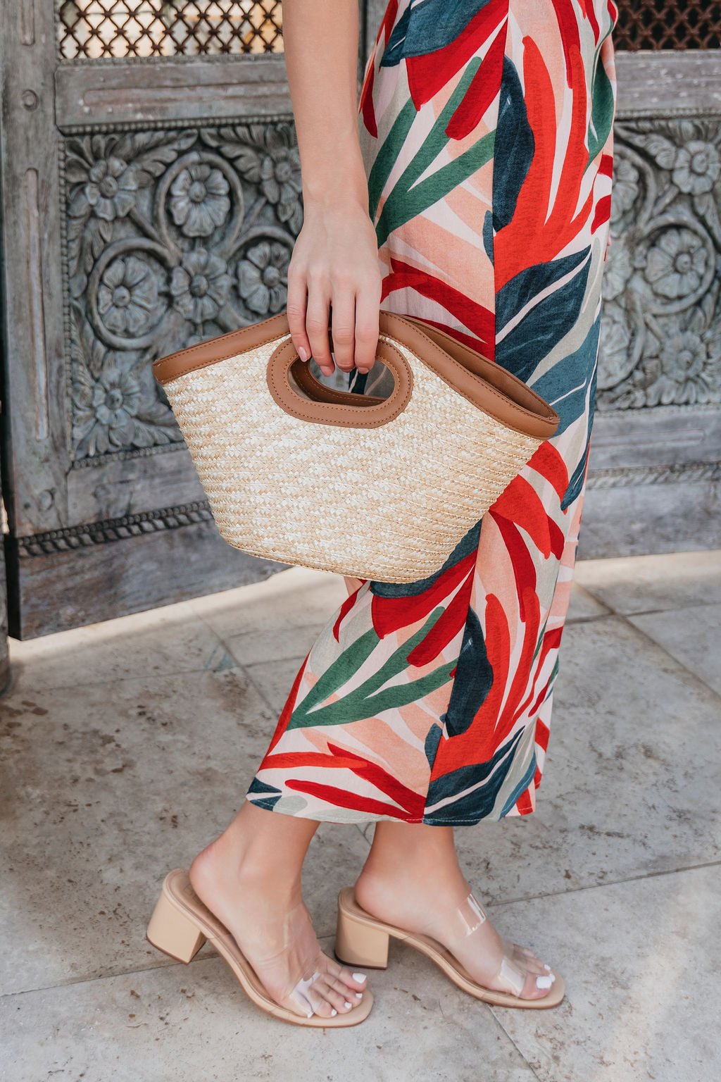 Woman in a colorful leaf dress holds the Beachwood Bliss Bucket Bag, styled with clear-strap heels on a stone floor.