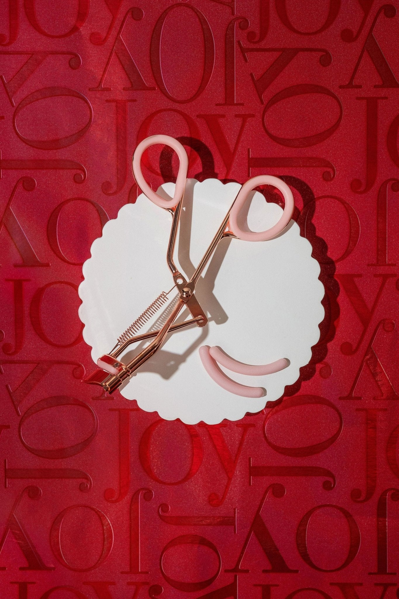 A white scalloped plate displays the Beauty Creations Pink Eyelash Curler and pads, arranged like a smile on a red Joy backdrop.