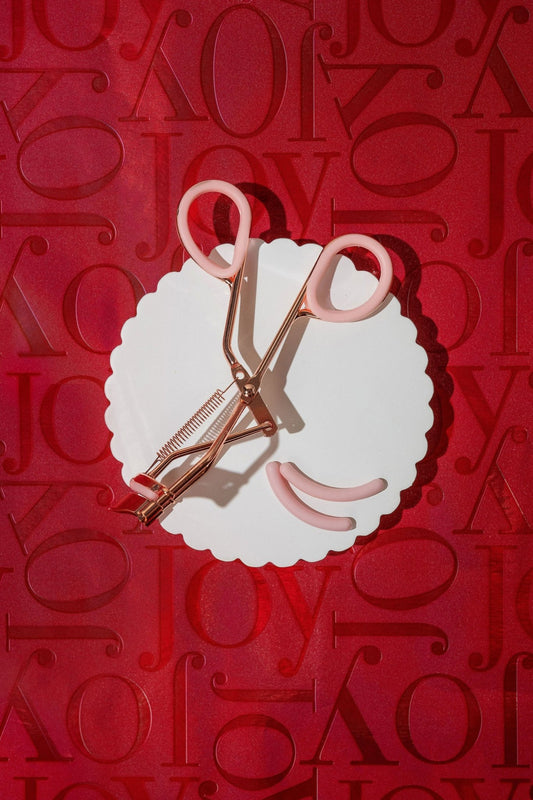 A white scalloped plate displays the Beauty Creations Pink Eyelash Curler and pads, arranged like a smile on a red Joy backdrop.