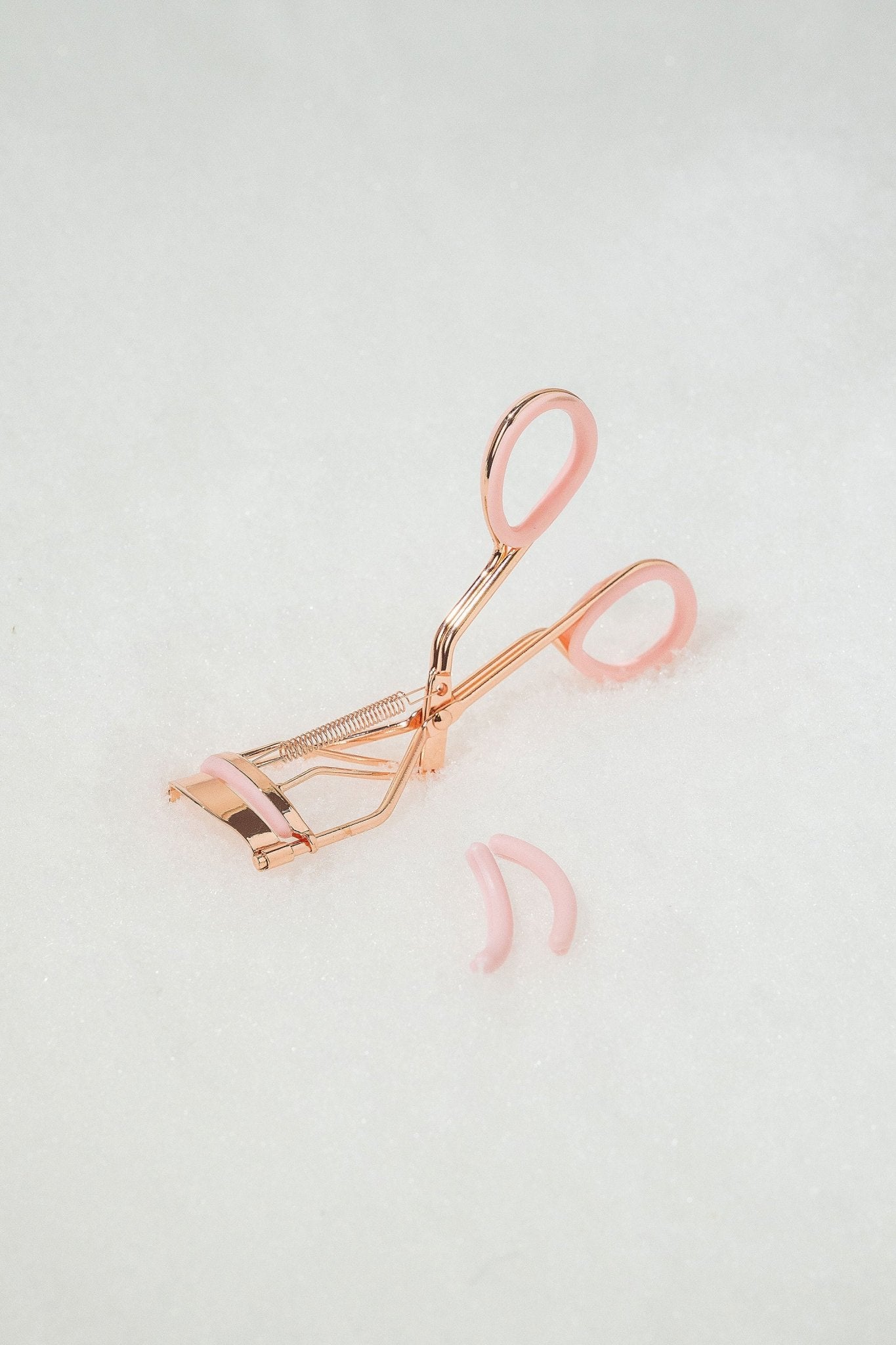 The Beauty Creations Pink Eyelash Curler with rose gold finish and pink handles is shown beside two pink silicone pads.