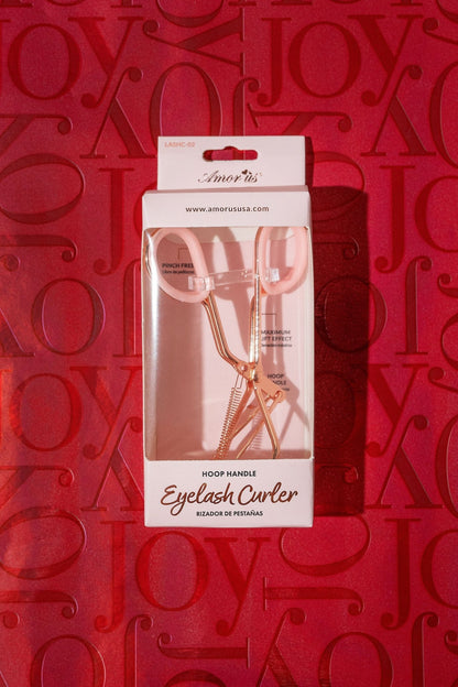 The Beauty Creations Pink Eyelash Curler with rose gold handles rests on a red background, "joy" embossed for an elevated routine.