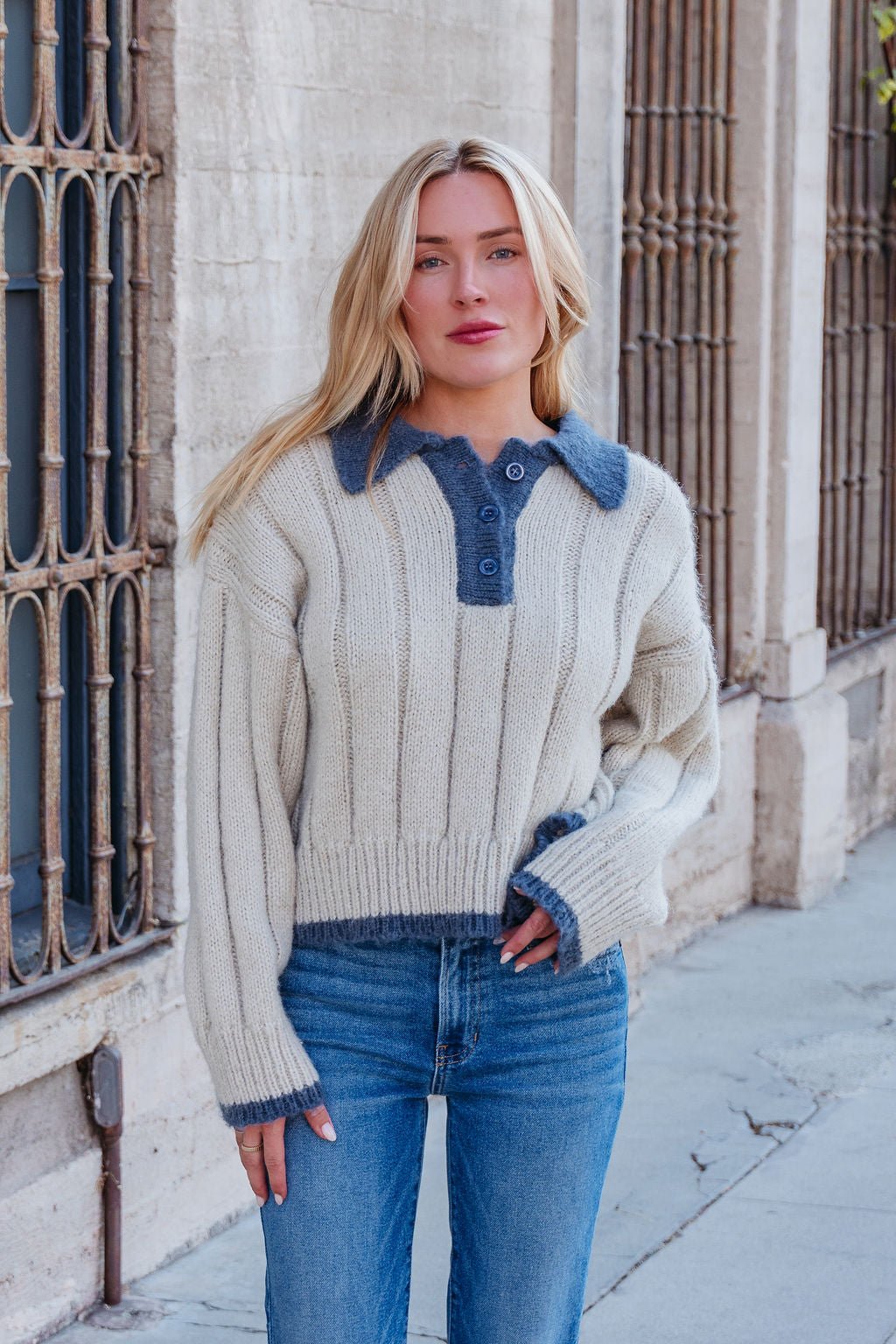 A woman models the Beige & Blue Button Up Sweater with blue jeans on a sidewalk in front of a beige building with tall windows.