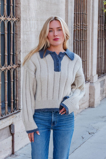 A woman models the Beige & Blue Button Up Sweater with blue jeans on a sidewalk in front of a beige building with tall windows.