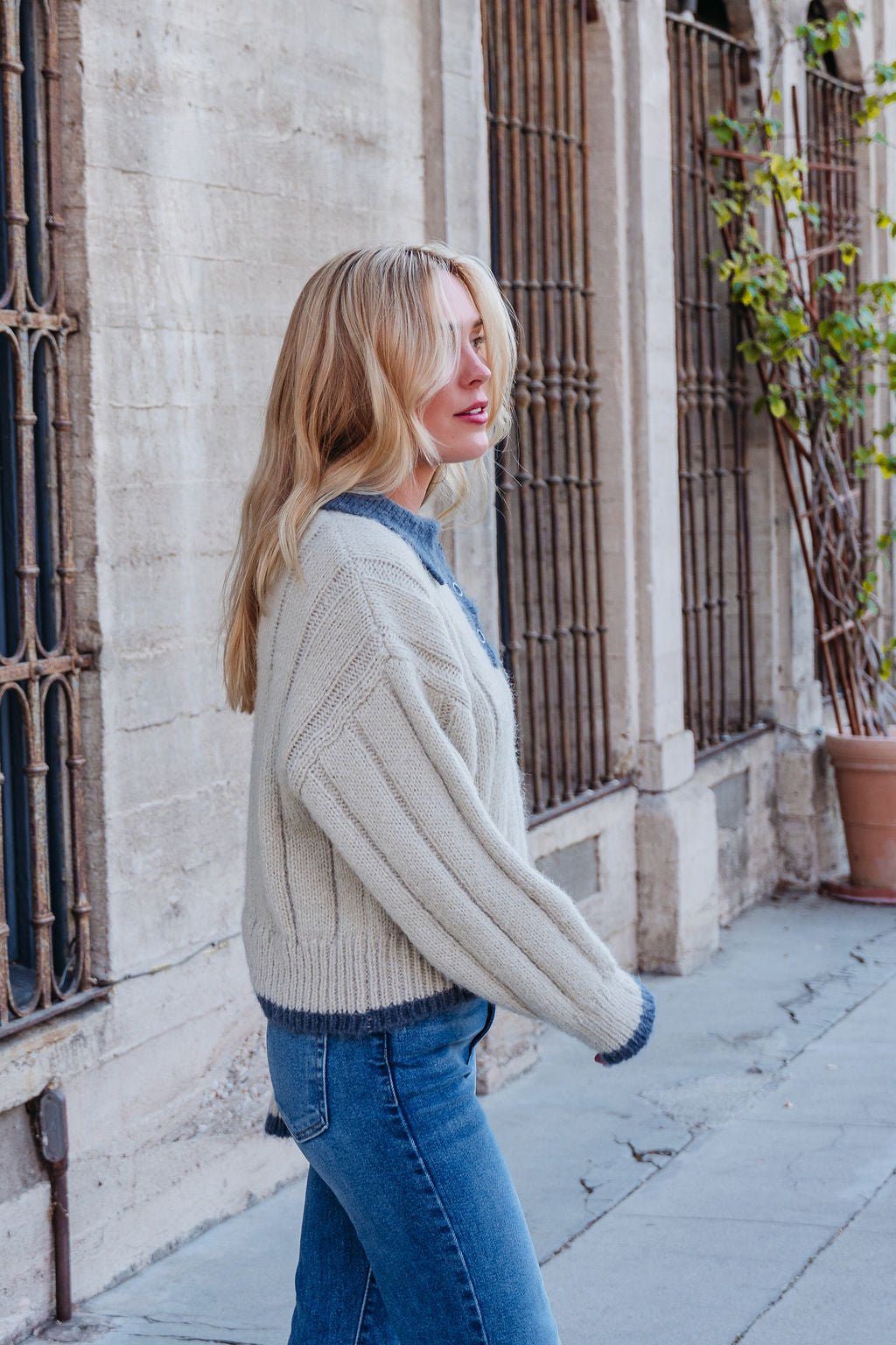 A woman with long blonde hair in a Beige & Blue Button Up Sweater and jeans walks beside a stone building.