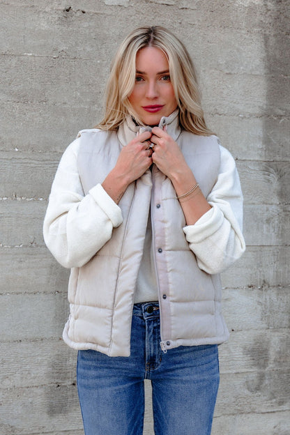 A woman with blonde hair wears a Beige Button Up Puffer Vest over a white sweater and blue jeans—ideal for fall layering.