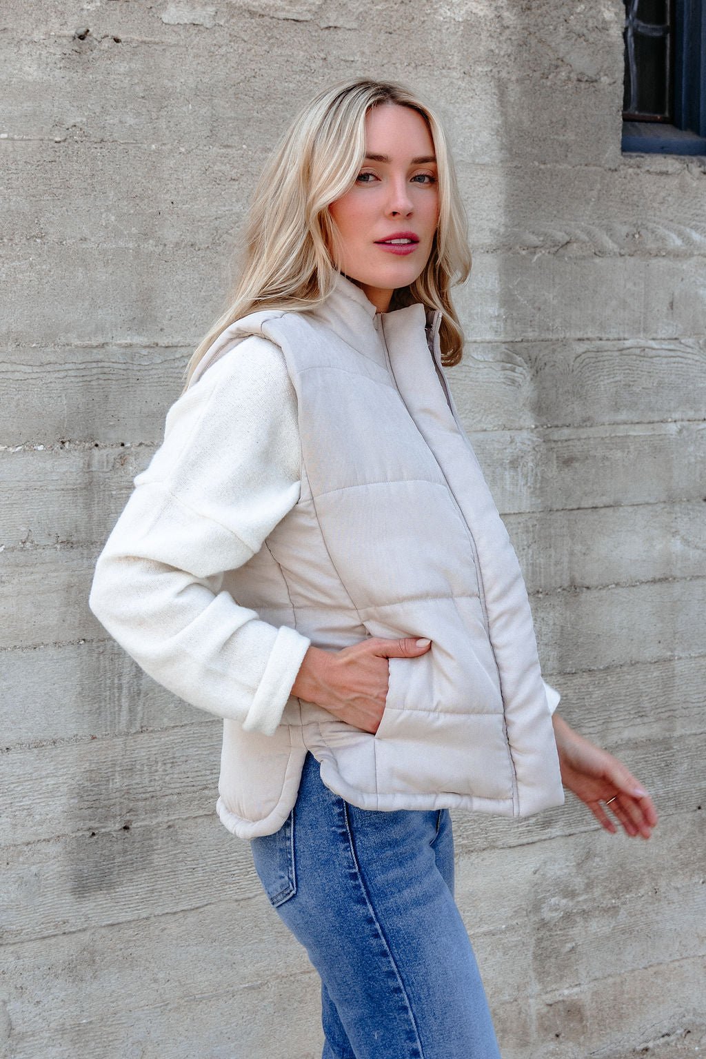 A woman in a Beige Button Up Puffer Vest and white sweater poses by a textured wall—ideal for chic fall layering.