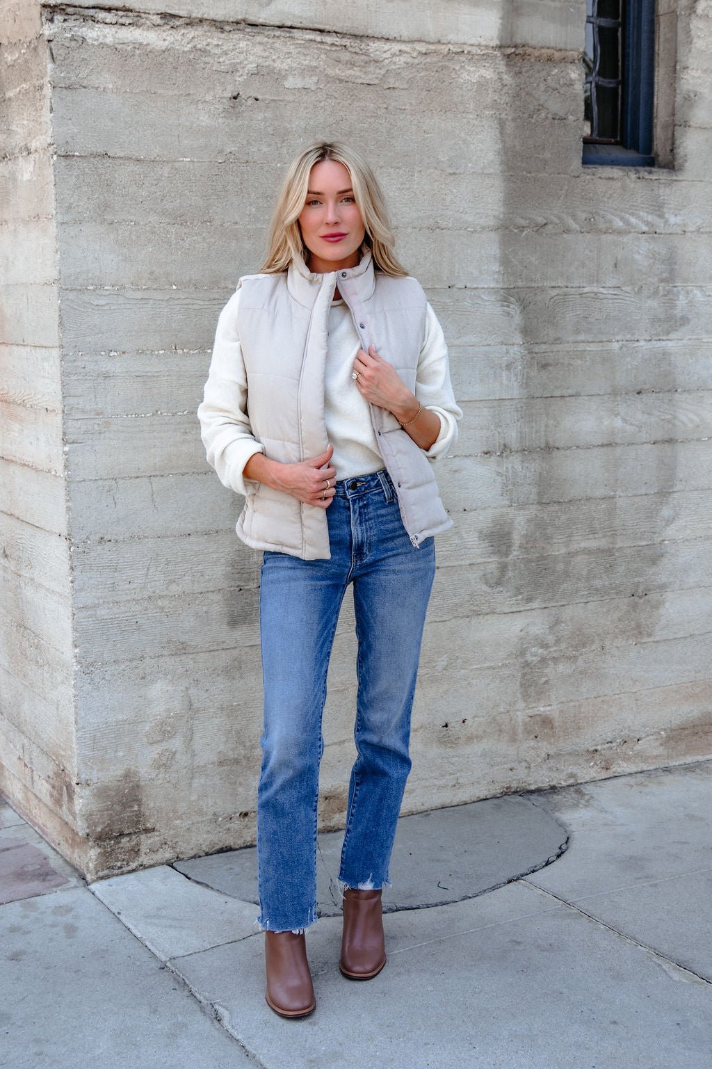 Woman in a Beige Button Up Puffer Vest, white sweater, blue jeans, and brown boots stands outside by a concrete wall.