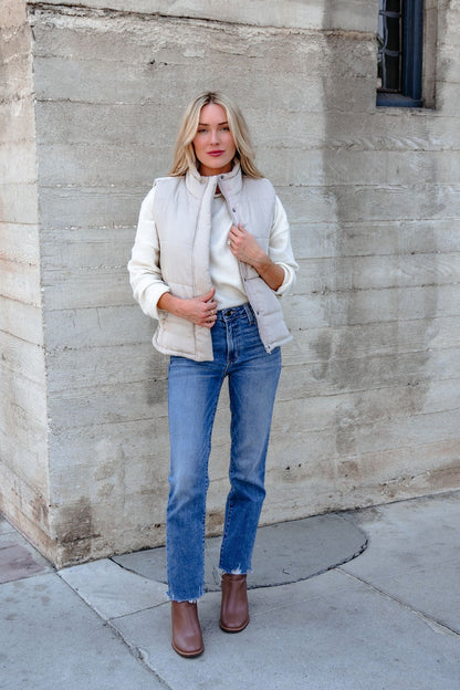 Woman in a Beige Button Up Puffer Vest, white sweater, blue jeans, and brown boots stands outside by a concrete wall.