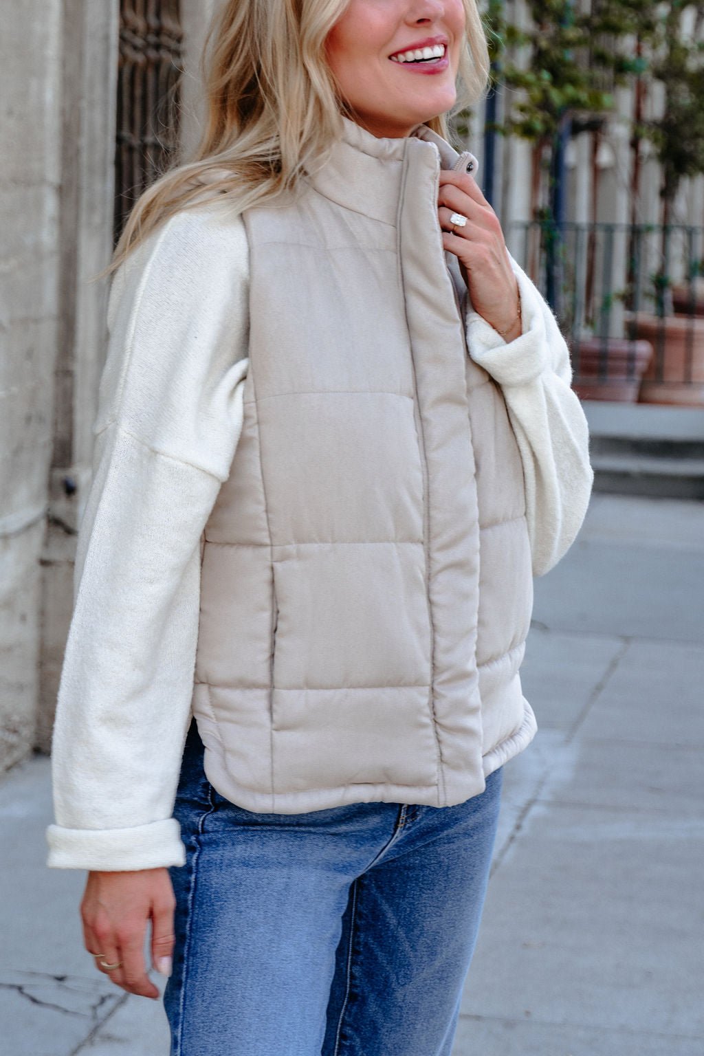 Woman smiling in a Beige Button Up Puffer Vest over a cream sweater and jeans—ideal for fall layering on a city sidewalk.