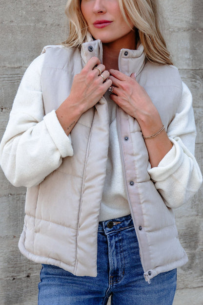 A woman in a Beige Button Up Puffer Vest, white sweater, and jeans stands by a concrete wall, effortlessly layering for fall.