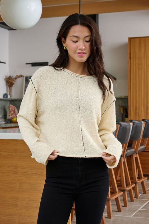 A woman with long dark hair wears the Beige Contrast Stitch Detail Sweater, standing in a modern wood-accented kitchen.