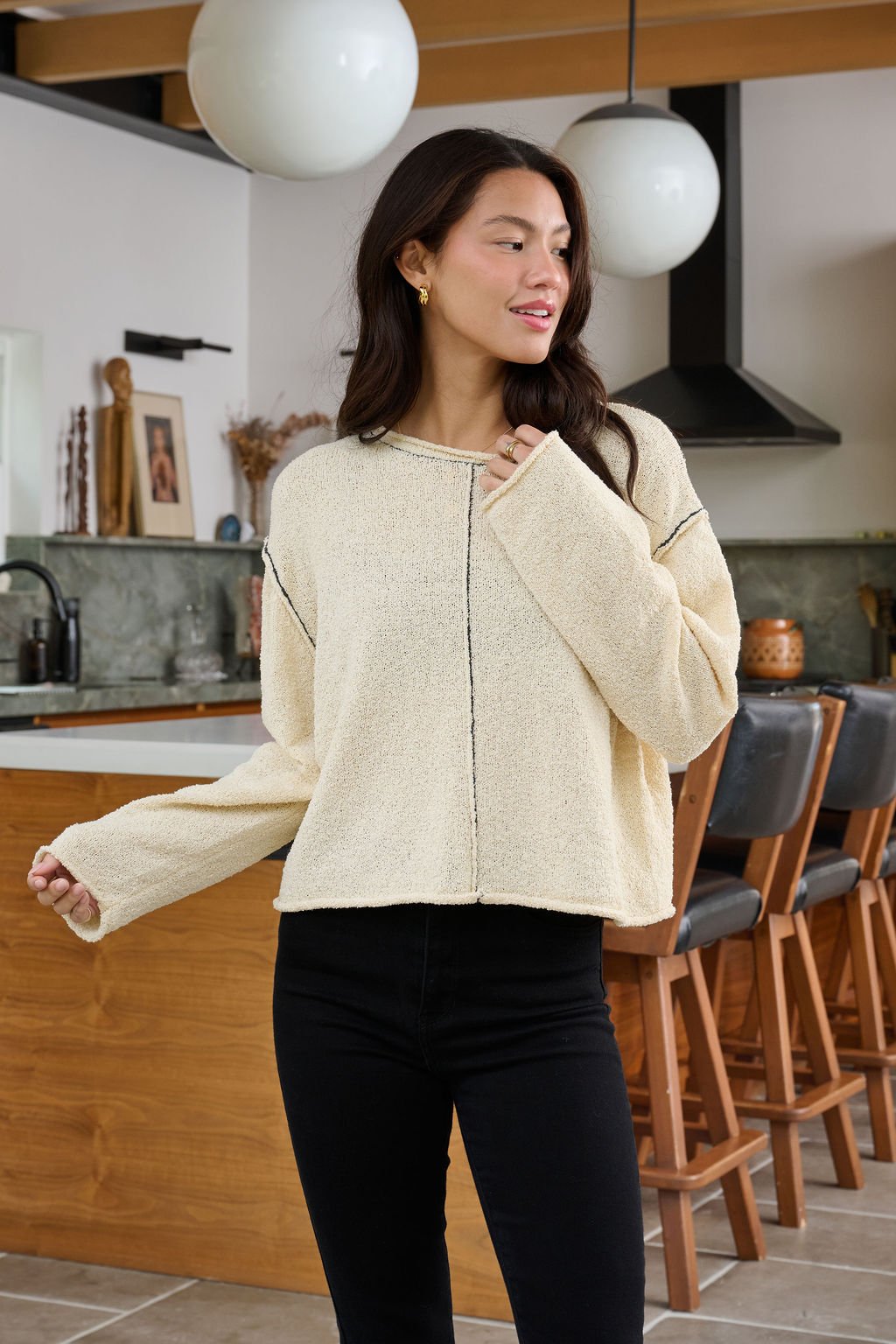 A woman wearing a Beige Contrast Stitch Detail Sweater and black jeans smiles and looks aside in a modern kitchen.